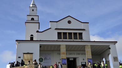 Gente de pie y sentada en los escalones de una iglesia blanca con campanario bajo un cielo azul despejado.. Abrir en galería a pantalla completa.